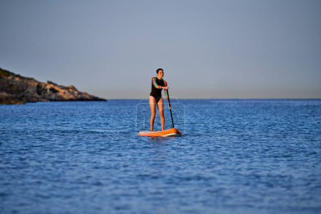 Photo for Full body of slim female surfer in swimwear standing on SUP board and rowing on rippling seawater during sunset and looking away - Royalty Free Image