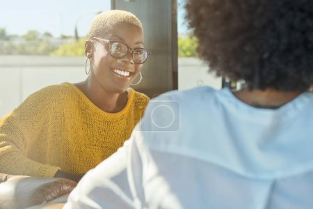 Photo for Delighted African American woman smiling while looking at friend sitting in cafe on sunny day - Royalty Free Image