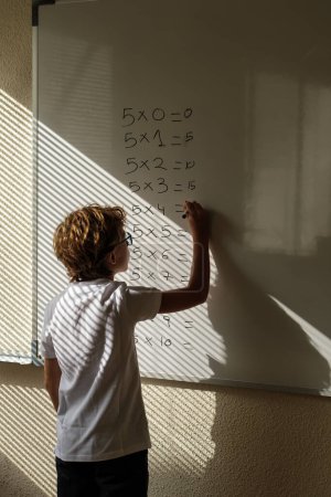 Photo for Back view of intelligent schoolboy standing writing multiplication table on whiteboard during math lesson in classroom - Royalty Free Image