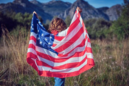 Photo for Smiling preteen boy in jeans overall standing in grassy meadow with raised arms with flag of United States of America - Royalty Free Image