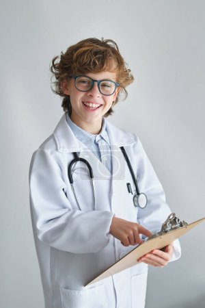 Foto de Niño alegre en bata médica con estetoscopio mostrando portapapeles mientras mira la cámara sobre fondo blanco - Imagen libre de derechos