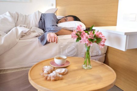 Téléchargez les photos : Femme adulte en pyjama dormant sur le lit près de la table en bois avec bouquet de fleurs et croissants au chevet dans la chambre à coucher à la maison - en image libre de droit