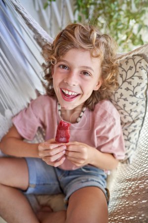 Foto de Niño feliz sonriendo ampliamente y comiendo sabroso helado paleta mientras descansa en la hamaca y mirando a la cámara - Imagen libre de derechos