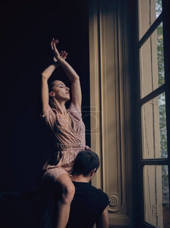 Photo for Charming ballerina with raised arms sitting on shoulder of unrecognizable strong male dancer near window of room with dim light - Royalty Free Image