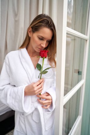 Téléchargez les photos : Femme réfléchie portant un peignoir éponge blanc sentant rose rouge tout en se tenant à la fenêtre de la suite de l'hôtel - en image libre de droit