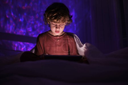 Photo for Focused boy in nightwear surfing tablet while sitting on bed in dark bedroom at night time at home - Royalty Free Image