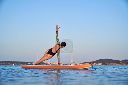 Téléchargez les photos : Vue de dos corps complet de femme mince anonyme en maillot de bain debout à Vasishthasana tout en pratiquant le yoga sur la planche SUP dans la mer calme en soirée - en image libre de droit