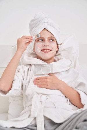 Photo for Positive child in bathrobe and towel on head sitting on bed and leaning on pillows while smearing moisturizing mask on face with brush - Royalty Free Image