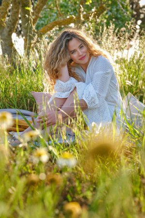 Foto de Sensible joven hembra vistiendo vestido blanco tocando el pelo ondulado y mirando a la cámara mientras está sentado en cuadros en el prado herboso en el soleado campo de verano - Imagen libre de derechos