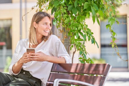 Photo for Smiling adult woman in casual clothes sitting on wooden bench with plant hanging from above and browsing cellphone on city street - Royalty Free Image