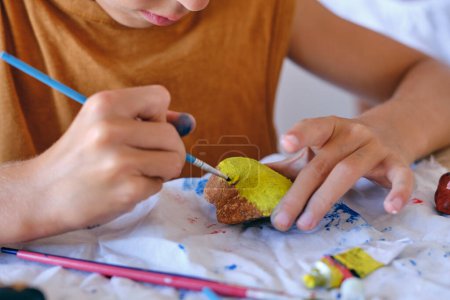 Photo for Crop anonymous preteen boy painting stone with watercolor while sitting at messy table and entertaining at weekend - Royalty Free Image
