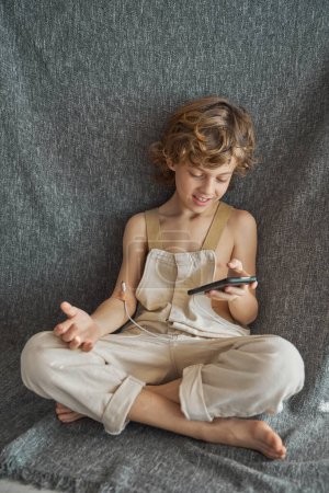 Foto de Cuerpo completo de niño adicto digital con cable de carga conectado al brazo con yeso adhesivo mirando a la pantalla del teléfono y sonriendo contra el fondo gris - Imagen libre de derechos