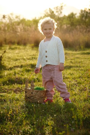 Foto de Contenido de cuerpo completo niña en ropa elegante con canasta de mimbre de pie en el prado herboso abundante en el día soleado y mirando a la cámara con bonita sonrisa - Imagen libre de derechos