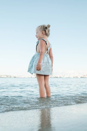 Photo for From below side view of happy little girl in summer dress standing in shallow sea on beach and looking away against cloudless sky - Royalty Free Image