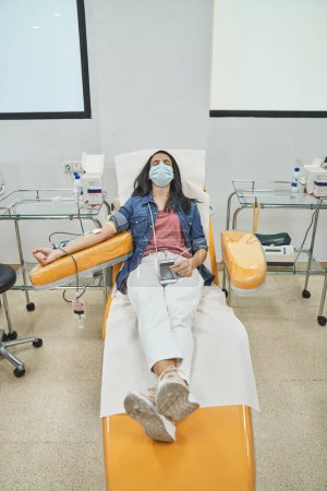 Téléchargez les photos : Corps complet de la femme adulte en vêtements décontractés et masque médical couché sur une chaise médicale avec les yeux fermés et donnant du sang avec un équipement spécial en laboratoire - en image libre de droit