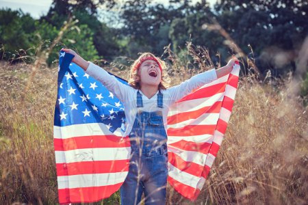 Foto de Niño contento cantando con bandera americana en las manos mientras se cierran los ojos en terreno herboso - Imagen libre de derechos