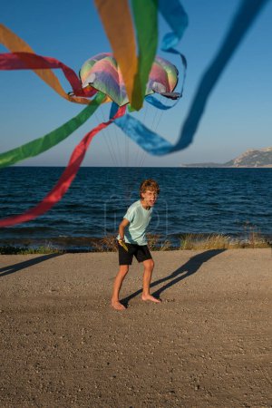 Téléchargez les photos : Corps complet de heureux énergique pieds nus garçon jouer avec multicolore cerf-volant flottant sur le vent tout en s'amusant pendant les vacances d'été sur le bord de la mer - en image libre de droit