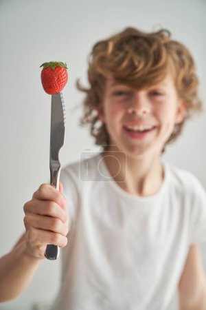 Foto de Niño despreocupado con baya dulce perforado con cuchillo sonriendo y mirando a la cámara sobre fondo blanco - Imagen libre de derechos