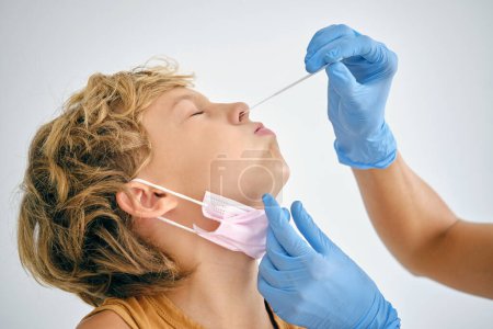 Foto de Médico con hisopo que toma la prueba del coronavirus de la nariz del niño con los ojos cerrados en la clínica sobre fondo claro - Imagen libre de derechos