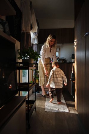Photo for Full body of mother and barefoot son in casual clothes standing on rug near wooden kitchen set in cozy dark room with daylight - Royalty Free Image