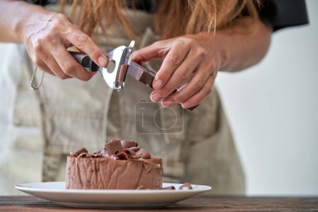 Photo for Crop female cook in apron with peeler garnishing delicious chocolate cake while chocolate shavings on top - Royalty Free Image