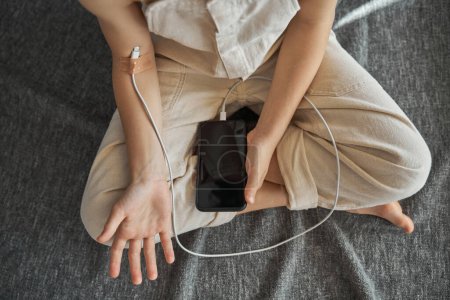 Photo for Top view of crop digital addicted child with charging cable connected to arm with adhesive plaster sitting on gray blanket with smartphone on legs - Royalty Free Image