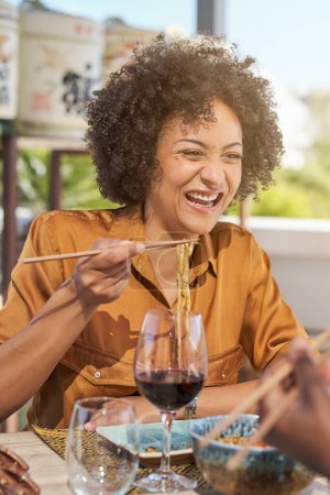 Téléchargez les photos : Heureuse ethnique bouclée femme en chemisier manger des nouilles avec des baguettes et boire du vin rouge sur la terrasse du restaurant - en image libre de droit