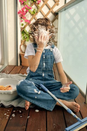 Photo for Barefoot anonymous preteen boy sitting on wooden balcony floor and hitting face with foam splashes while washing glass in sunlight - Royalty Free Image