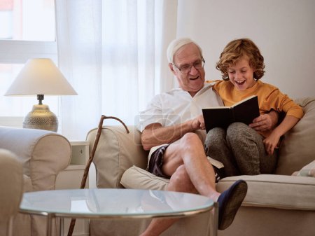 Téléchargez les photos : Homme âgé attentionné avec un petit-fils positif lisant un livre intéressant tout en étant assis sur un canapé confortable dans le salon lumineux à la maison - en image libre de droit