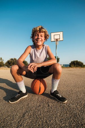 Téléchargez les photos : Corps complet de gai garçon préadolescent en vêtements de sport assis sur le basket-ball à l'aire de jeux et regardant la caméra le jour ensoleillé - en image libre de droit
