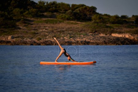 Téléchargez les photos : Vue latérale d'une athlète féminine flexible en maillot de bain prenant la pose Adho Mukha Svanasana avec jambe relevée sur la planche SUP tout en pratiquant le yoga en mer - en image libre de droit