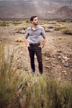 Foto de Joven positivo en traje de moda de pie en suelo pedregoso cerca de las montañas y mirando hacia otro lado - Imagen libre de derechos