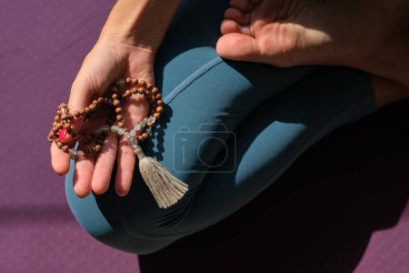Téléchargez les photos : Vue du dessus de la récolte anonyme pieds nus femelle avec chapelet pratiquant le yoga à Padmasana tout en étant assis sur le tapis le jour ensoleillé - en image libre de droit