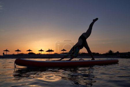 Photo for Full body of young woman in swimsuit taking Adho Mukha Shvanasana pose with leg raised while practicing yoga on SUP board on sundown - Royalty Free Image