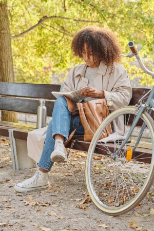 Téléchargez les photos : Corps complet de jeune femme afro-américaine en tenue décontractée avec bouclé livre de lecture de cheveux sur banc en bois dans le parc près de vélo - en image libre de droit