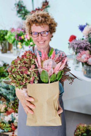 Foto de Mujer alegre de mediana edad en gafas sonriendo y mirando a la cámara mientras trabaja en la tienda de flores y demostrando ramo de flores arreglado - Imagen libre de derechos