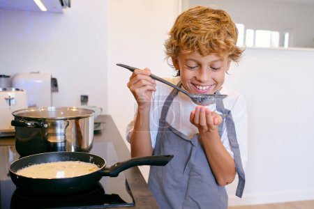 Téléchargez les photos : Contenu enfant dans tablier avec sauce blanche dans spatule cuillère debout près de la poêle sur la cuisinière à la maison - en image libre de droit