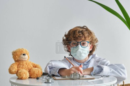 Foto de Niño en anteojos y máscara respiratoria mirando a la cámara mientras está sentado en la mesa con oso suave y portapapeles sobre fondo blanco - Imagen libre de derechos