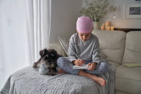 Téléchargez les photos : Corps entier d'un garçon pieds nus concentré portant un pyjama et un bandana rose à l'appui des patientes atteintes du cancer du sein assis près d'un chien miniature Schnauzer et écrivant dans un carnet dans le salon - en image libre de droit