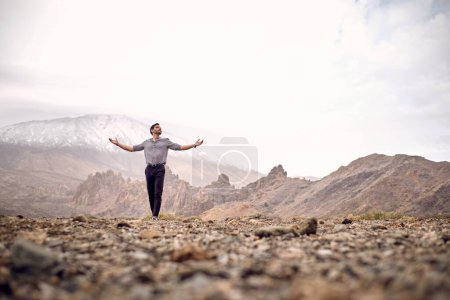 Téléchargez les photos : Terrain plein niveau de touriste hispanique mâle debout avec les bras tendus sur un sol rocheux sec contre la montagne Teide et regardant loin dans les îles Canaries de Tenerife Espagne - en image libre de droit