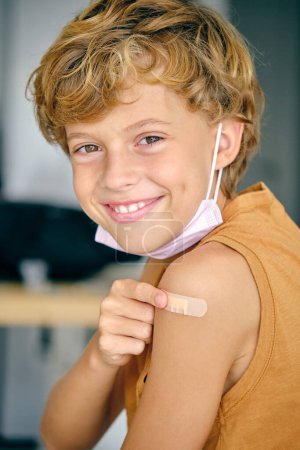 Photo for Side view of content child with brown hair and sterile mask looking at camera while applying medical patch on shoulder - Royalty Free Image