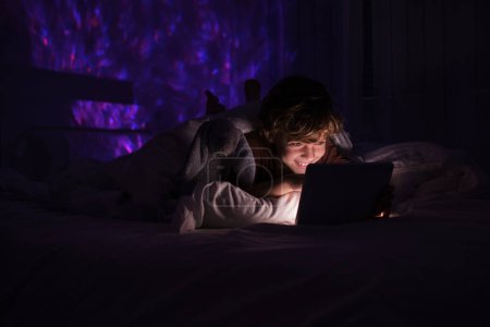 Téléchargez les photos : Joyeux garçon naviguant tablette moderne tout en étant couché sur le lit sous la couverture dans la chambre sombre avec des lumières incandescentes la nuit - en image libre de droit