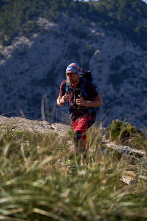 Foto de Viajero masculino activo con mochila trekking en ladera de montaña cubierta de hierba durante la aventura de verano contra acantilado rocoso - Imagen libre de derechos