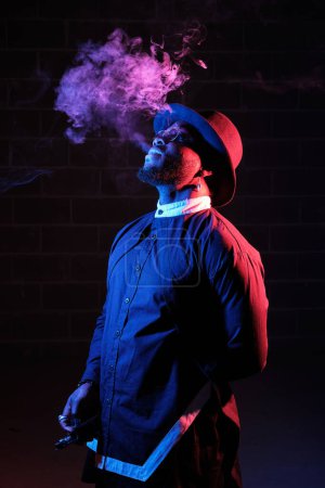 Photo for Side view of ethnic guy in stylish outfit and hat standing against black background and exhaling fume in neon illumination - Royalty Free Image