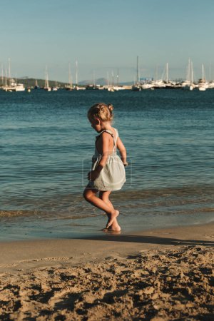 Téléchargez les photos : Vue latérale de gamin ludique en robe bleue marchant sur la plage de sable près de la mer ondulante avec des yachts au crépuscule - en image libre de droit