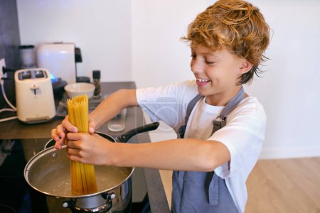 Téléchargez les photos : Contenu enfant mettre des pâtes non cuites dans une casserole avec de l'eau bouillante sur plaque de cuisson dans la cuisine de la maison - en image libre de droit