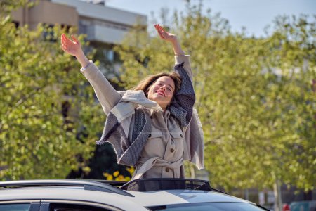 Téléchargez les photos : Joyeux jeune femme en manteau debout avec les yeux fermés et les bras levés dans le toit ouvrant de la voiture dans le jour ensoleillé du printemps - en image libre de droit