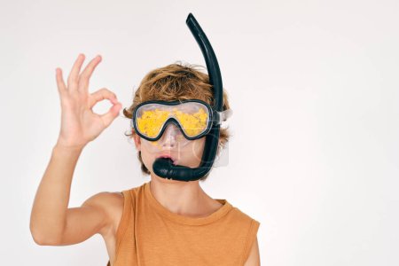 Foto de Niño en máscara de buceo con snorkel y algas amarillas demostrando gesto OK sobre fondo claro - Imagen libre de derechos