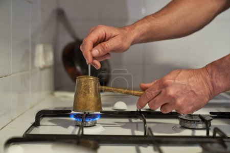 Foto de Cultivo hombre anónimo verter café aromático caliente fresco en cezve en la estufa de gas en la cocina mientras se agita con una cucharadita - Imagen libre de derechos