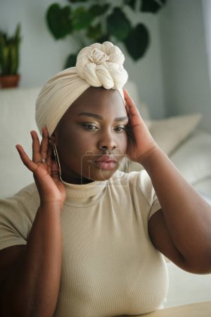 Foto de Encantadora mujer negra con maquillaje ajustando el turbante tradicional en la cabeza sobre un fondo borroso dormitorio - Imagen libre de derechos